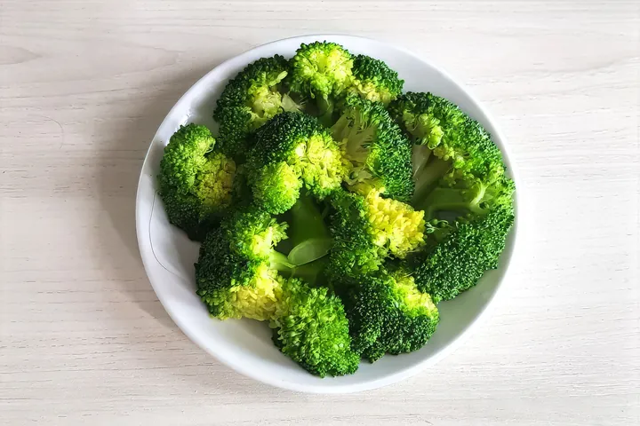 A white bowl of steamed broccoli florets on a pale wooden table