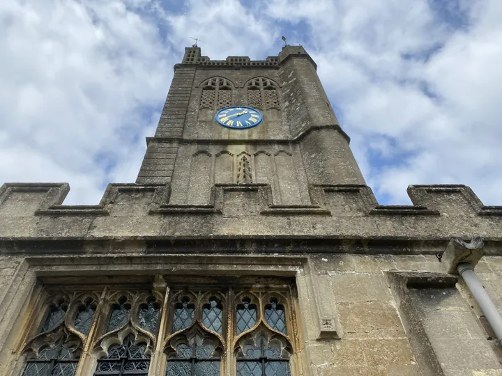 Looking up at a stone church tower with a blue clock face under a cloudy sky