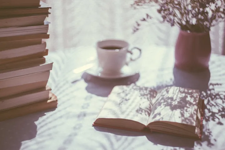 A cup of coffee and an open book on a table in dappled window light