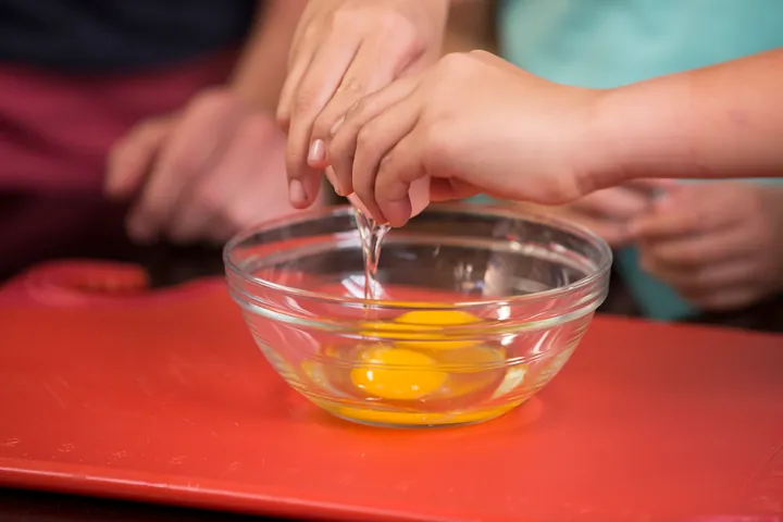 Hands cracking an egg into a glass bowl on a red chopping board