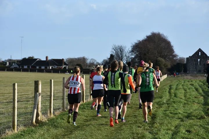 A group of club runners seen from behind on a grassy cross-country course beside a wooden fence