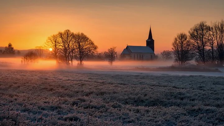 A frosted field at sunrise, with low mist around a small country church and bare trees