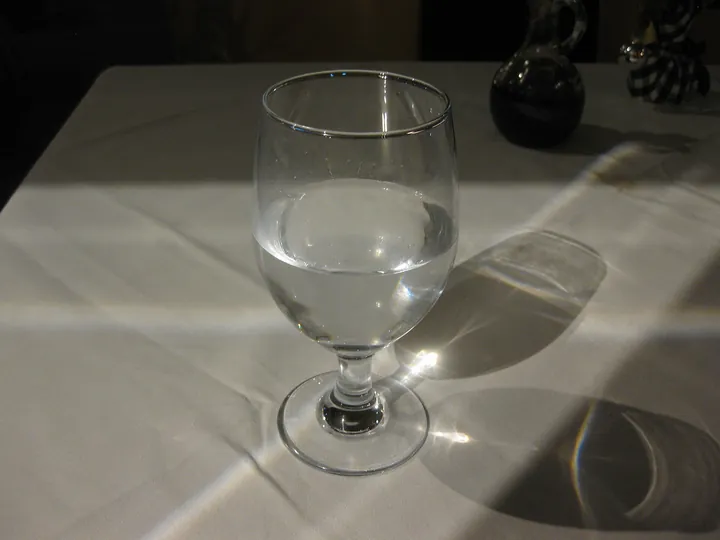 A glass of water on a white tablecloth, sunlight casting its shadow across the table