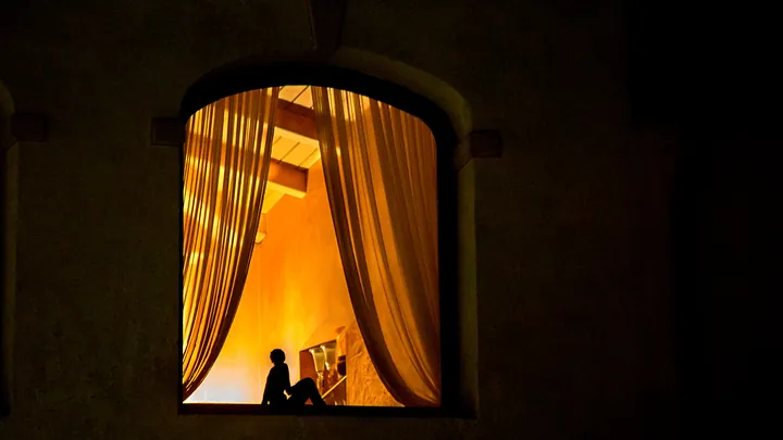 A warmly lit window at night, curtains drawn back and a figure sitting in silhouette inside