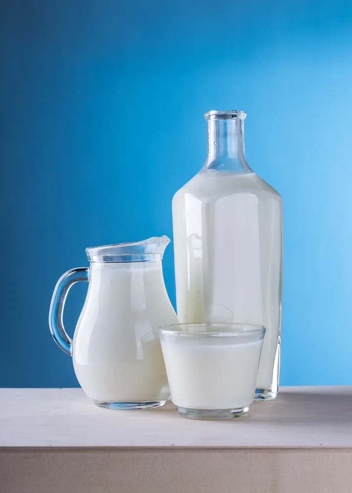 A jug, a bottle and a glass of milk on a white shelf against a blue wall
