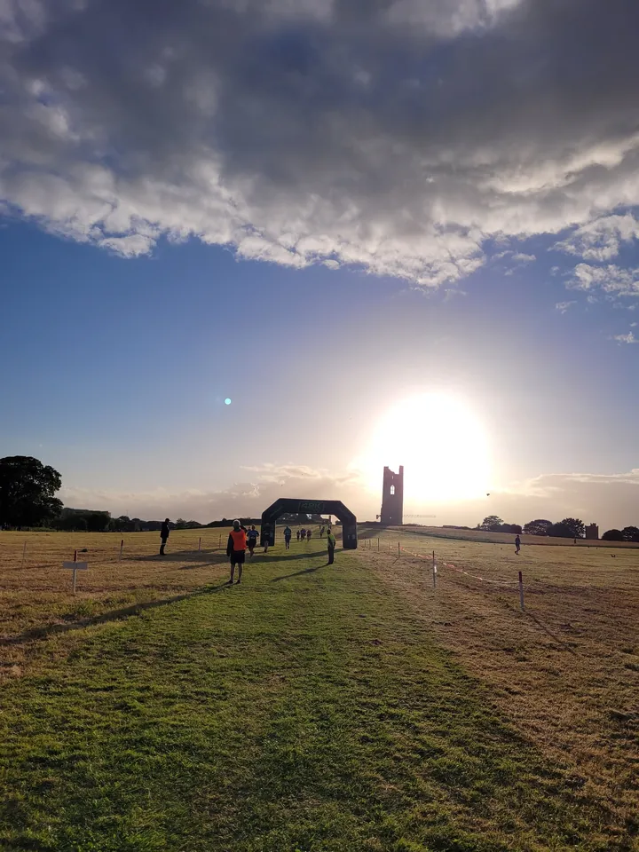 Distant runners crossing an open field towards a low winter sun, with a ruined tower on the horizon